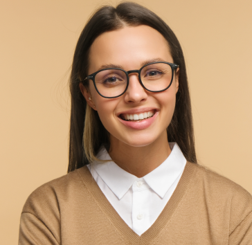 Una donna con gli occhiali sorride alla telecamera, indossando una camicia bianca con colletto e un maglione beige, su uno sfondo beige.