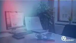 Scrivania da ufficio con monitor che mostra grafici, documenti aperti, tastiera, mouse, due tazze da caffè e una pianta. Una finestra sullo sfondo mostra una vista esterna.